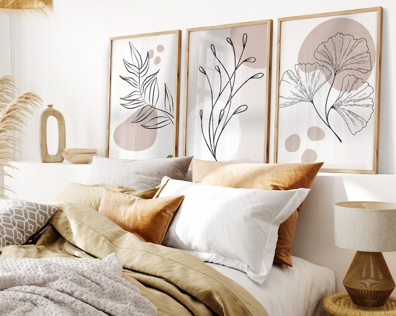 Könnte beinhalten: Drei gerahmte Drucke mit minimalistischen Linienzeichnungen von Blättern und Zweigen auf hellem Hintergrund mit dezenten rosafarbenen Akzenten. Die Drucke hängen an einer weißen Wand über einem Bett mit brauner Bettwäsche und Kissen.