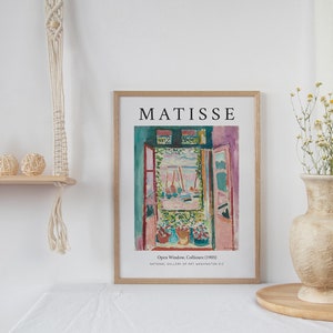 May include: A framed print of a painting by Henri Matisse titled "Open Window, Collioure (1905)". The painting depicts a view from a window with a bright blue sky, boats, and a lush green garden. The frame is wooden and has a white mat.