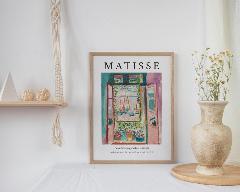 May include: A framed print of a painting by Henri Matisse titled "Open Window, Collioure (1905)". The painting depicts a view from a window with a bright blue sky, boats, and a lush green garden. The frame is wooden and has a white mat.