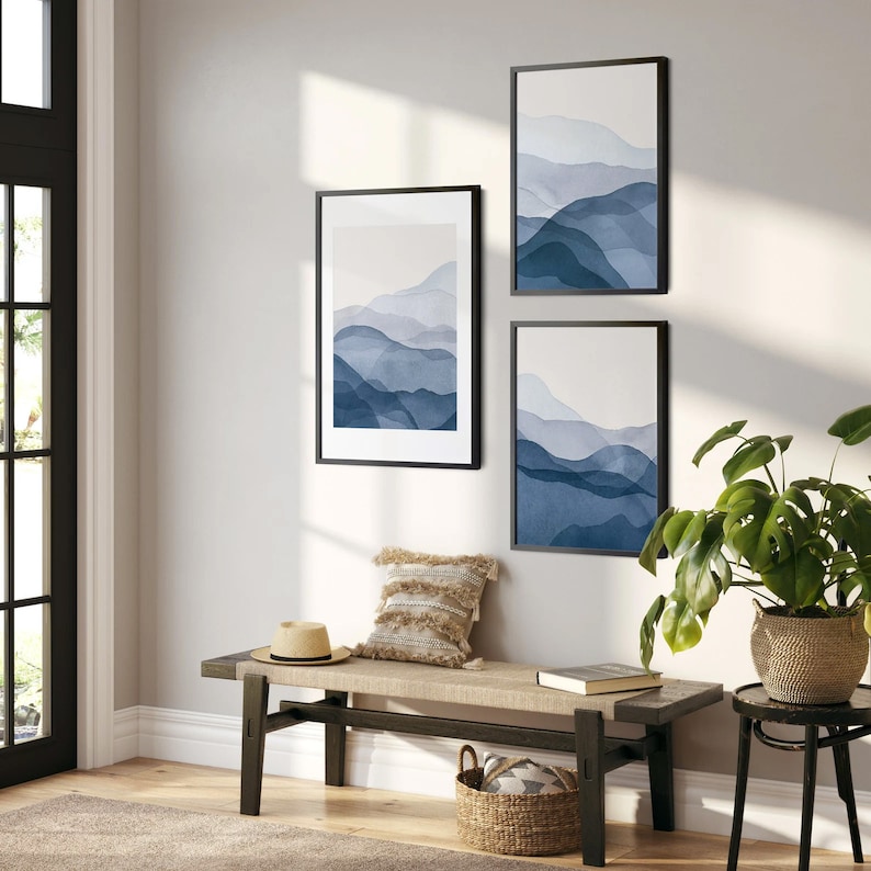 May include: Three framed abstract watercolor prints of blue mountains on a white background, hanging on a white wall above a wooden bench with a woven basket and a potted plant.