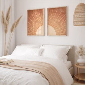 Op de afbeelding: Een wit bed met een beige deken en twee witte kussens. Twee ingelijste prints van een zonnestralenontwerp in oranje en bruine tinten hangen boven het bed.