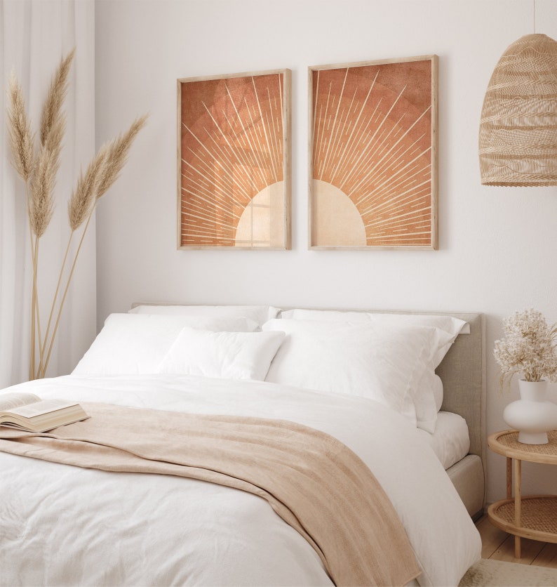Op de afbeelding: Een wit bed met een beige deken en twee witte kussens. Twee ingelijste prints van een zonnestralenontwerp in oranje en bruine tinten hangen boven het bed.