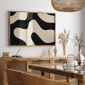 May include: A black and white abstract art print with a beige background hangs on a white wall above a wooden cabinet with woven rattan doors. The cabinet is in a dining room with a wooden table and chairs.