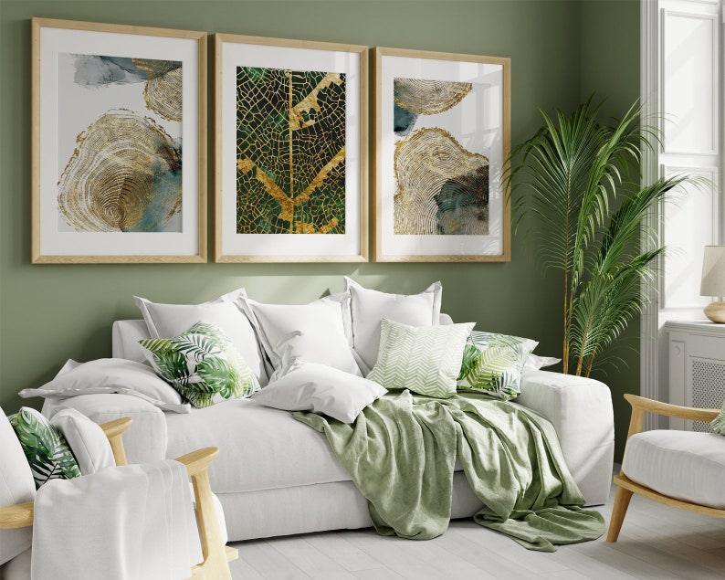 Puede incluir: Un sof&aacute; blanco con mantas y almohadas verdes con un patr&oacute;n de hojas tropicales. Tres impresiones de arte abstracto enmarcadas con acentos dorados y verdes cuelgan de una pared verde salvia detr&aacute;s del sof&aacute;.