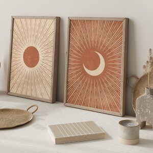 Puede incluir: Dos impresiones enmarcadas con un dise&ntilde;o minimalista que presenta un sol y una luna creciente en color terracota con l&iacute;neas blancas que irradian hacia afuera. Las impresiones se colocan contra una pared blanca con una cesta de mimbre y un jarr&oacute;n de cer&aacute;mica.
