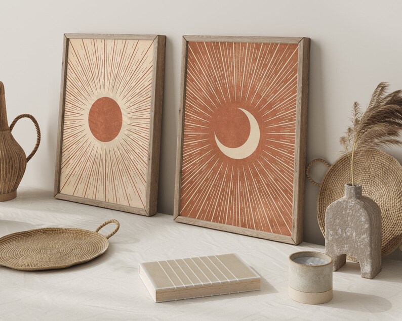 Puede incluir: Dos impresiones enmarcadas con un dise&ntilde;o minimalista que presenta un sol y una luna creciente en color terracota con l&iacute;neas blancas que irradian hacia afuera. Las impresiones se colocan contra una pared blanca con una cesta de mimbre y un jarr&oacute;n de cer&aacute;mica.