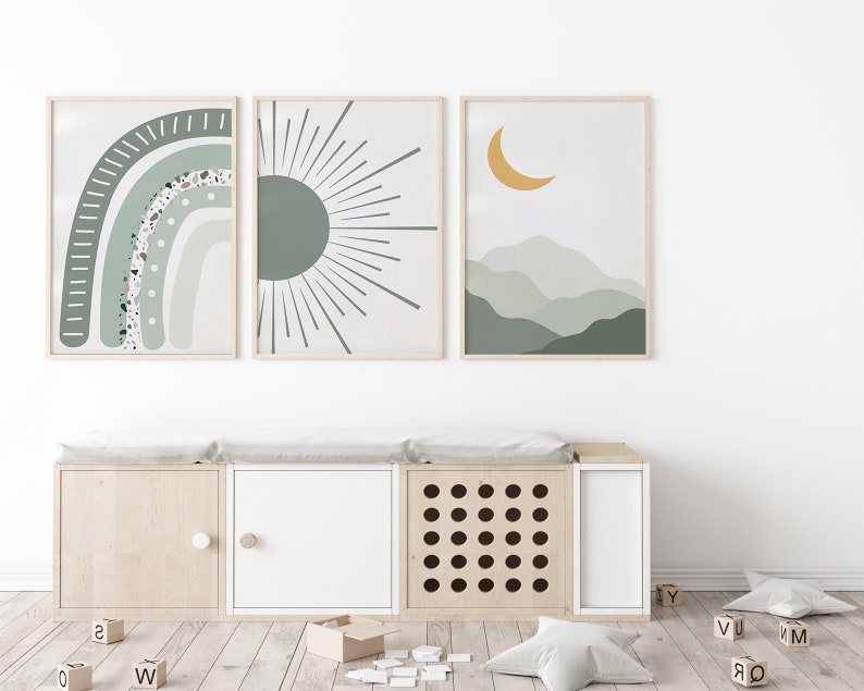 May include: Three framed prints with abstract designs in shades of green and yellow. The prints feature a rainbow, a sun, and a crescent moon over a mountain range. The prints are hanging on a white wall above a wooden storage bench with three compartments.