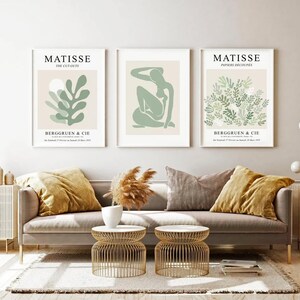 May include: Three framed art prints with a white background and green and beige abstract designs. The prints are titled "Matisse The Cut-Outs", "Matisse Papiers Decoup&eacute;s", and "Matisse Papiers Decoup&eacute;s". The prints are hanging on a white wall above a gray couch with two throw pillows.