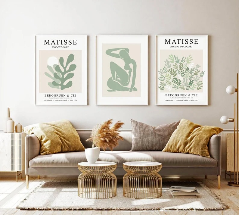 May include: Three framed art prints with a white background and green and beige abstract designs. The prints are titled "Matisse The Cut-Outs", "Matisse Papiers Decoup&eacute;s", and "Matisse Papiers Decoup&eacute;s". The prints are hanging on a white wall above a gray couch with two throw pillows.