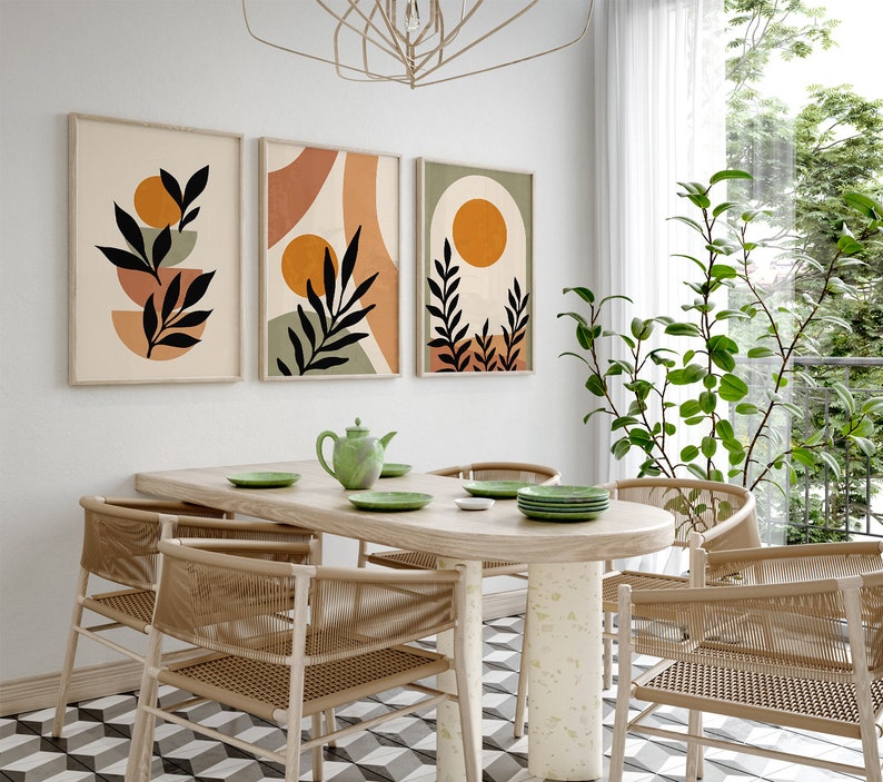 Op de afbeelding: Een eetkamer met een tafel van licht hout en zes stoelen. De stoelen zijn gemaakt van gevlochten riet en hebben een natuurlijke bruine kleur. Er hangen drie abstracte kunstprints aan de muur achter de tafel. De prints tonen geometrische vormen en bladeren in tinten bruin, oranje en groen.