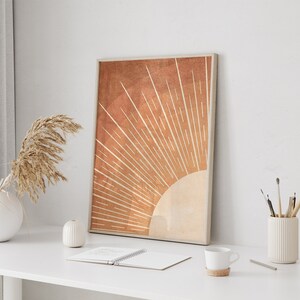 Op de afbeelding: Een minimalistische kunstprint met een gestileerde zon in oranje en beige tinten. De zon heeft een stralend patroon van lijnen die vanuit het midden uitstralen. De print is ingelijst en wordt getoond op een wit bureau met andere decoraties.