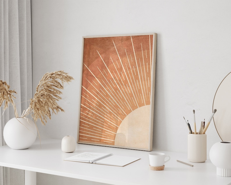 Op de afbeelding: Een minimalistische kunstprint met een gestileerde zon in oranje en beige tinten. De zon heeft een stralend patroon van lijnen die vanuit het midden uitstralen. De print is ingelijst en wordt getoond op een wit bureau met andere decoraties.