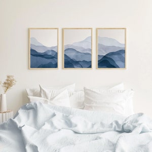 May include: Three framed watercolor prints of abstract blue mountains. The prints are hung on a white wall above a white bed with white bedding.