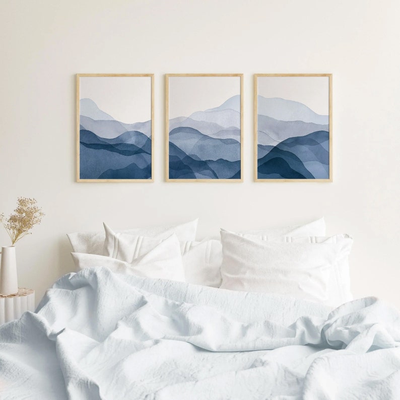 May include: Three framed watercolor prints of abstract blue mountains. The prints are hung on a white wall above a white bed with white bedding.