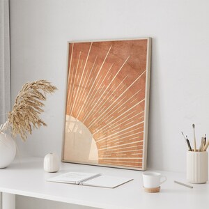 Op de afbeelding: Een ingelijste print van een minimalistisch zonnestralenontwerp in tinten oranje en bruin. De zonnestralen zijn gecentreerd op een witte cirkel en hebben een textuurrijke achtergrond.
