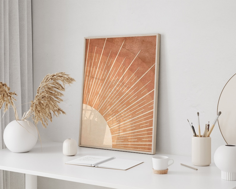 Op de afbeelding: Een ingelijste print van een minimalistisch zonnestralenontwerp in tinten oranje en bruin. De zonnestralen zijn gecentreerd op een witte cirkel en hebben een textuurrijke achtergrond.