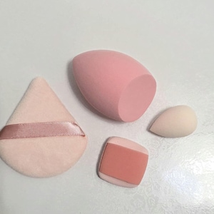 Op de afbeelding: Een verzameling make-up applicators in verschillende vormen en maten. Inbegrepen zijn een roze druppelvormige spons, een lichtroze poederdons met een lint, een kleine beige spons en een roze en beige rechthoekige spons.