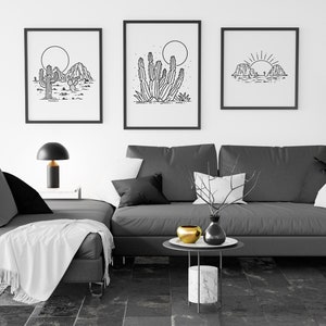 May include: Three black and white line art prints of desert landscapes with cacti and mountains. The prints are framed and hung on a white wall above a gray sectional sofa with white throw pillows and a white throw blanket.