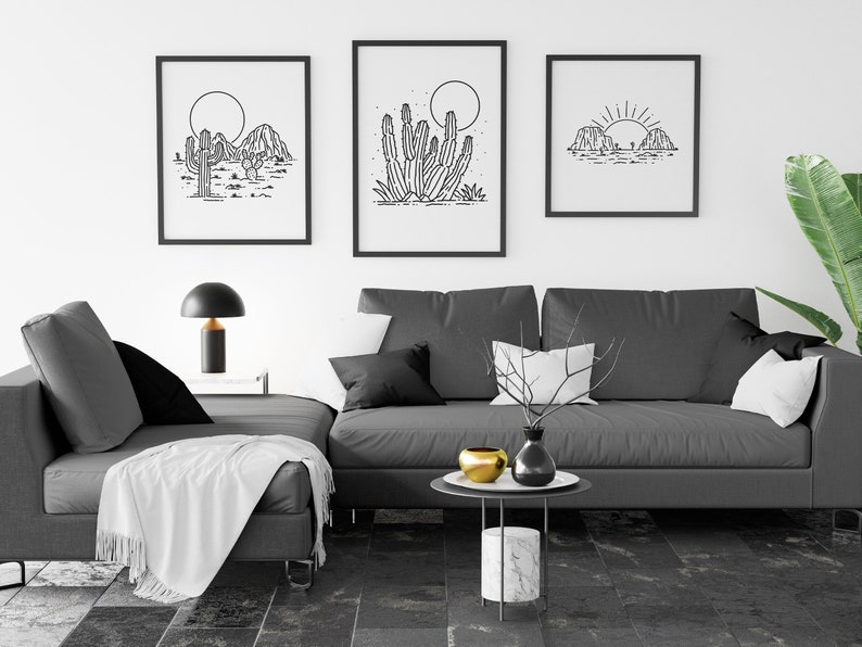 May include: Three black and white line art prints of desert landscapes with cacti and mountains. The prints are framed and hung on a white wall above a gray sectional sofa with white throw pillows and a white throw blanket.