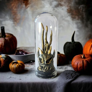 Peut inclure: Une cloche en verre contenant une figurine de créature dorée et noire. La figurine a une expression menaçante et semble être faite de résine. La cloche est posée sur une table avec des citrouilles et des courges autour.