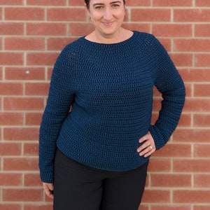 Puede incluir: Una mujer lleva un suéter de crochet azul oscuro con mangas largas y cuello redondo. Está de pie frente a una pared de ladrillo.