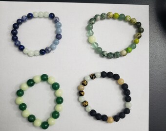 May include: Four bracelets made of different colored beads. The bracelets are arranged in a square pattern on a white surface. The bracelets are made of blue, green, black, and white beads.