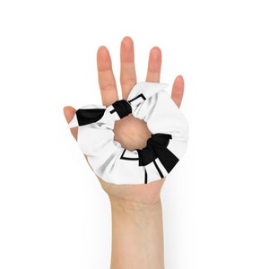 May include: A white scrunchie with a black geometric design. The scrunchie is tied in a knot and is being held in a hand.