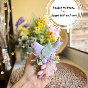 Peut inclure: Un oiseau en tissu lavande avec une feuille verte et des ornements floraux. L'oiseau a un bec noir et est orné d'un ruban de dentelle et de petits œufs décoratifs. Le texte sur l'image indique "Sewing pattern + videos instructions."