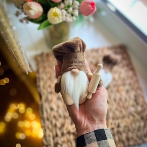 Könnte beinhalten: Eine braune und beige Zwergenpuppe mit Kochmütze, die ein kleines hölzernes Nudelholz hält. Der Zwerg hat einen langen weißen Bart und einen beigefarbenen Körper. Die Puppe befindet sich vor einem verschwommenen Hintergrund mit Blumen.