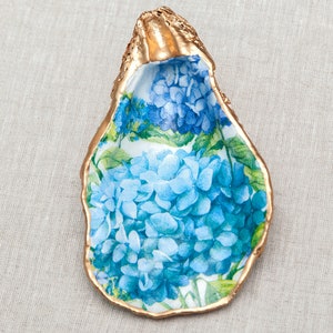May include: A gold-rimmed oyster shell dish with a blue and green floral pattern. The dish is decorated with a design of blue hydrangeas.