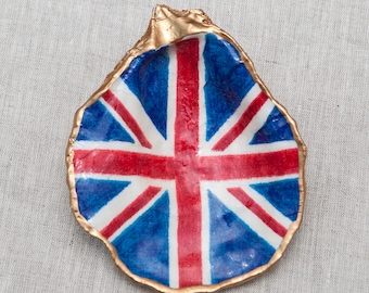 Puede incluir: Una concha de ostra con un borde dorado, pintada con la bandera de la Union Jack. La bandera es roja, blanca y azul.