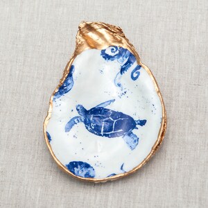 May include: A white oyster shell with a gold rim, painted with a blue sea turtle design.