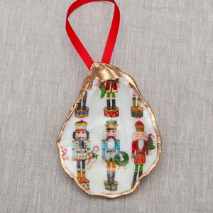 May include: A white oyster shell Christmas ornament with a gold trim. The shell is decorated with a pattern of nutcracker figures in various colors.