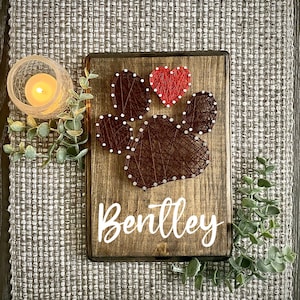 May include: A wooden sign with a brown paw print made of string and a red heart. The name "Bentley" is written in white lettering below the paw print.