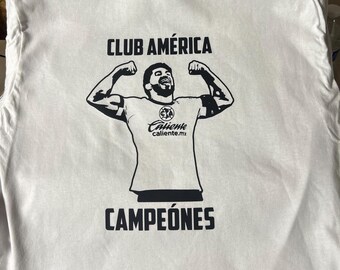 Puede incluir: Camiseta blanca con un gráfico negro de una figura flexionando los brazos, con el texto "CLUB AMÉRICA" arriba y "CAMPEONES" abajo. La figura lleva una camiseta con el logo "Caliente caliente.mx".