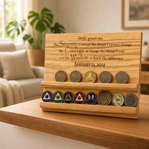 May include: A light-colored wooden display stand featuring two horizontal grooves holding several circular metal sobriety coins. The wood is engraved with the Serenity Prayer and the date JANUARY 11, 2014. The coins vary in color and design.