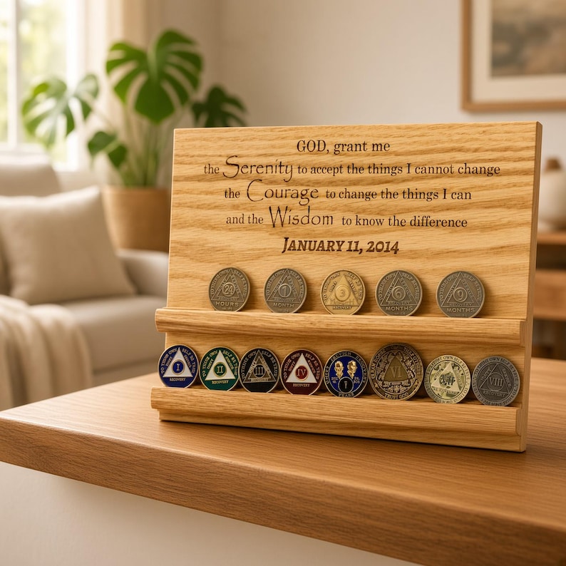 May include: A light-colored wooden display stand featuring two horizontal grooves holding several circular metal sobriety coins. The wood is engraved with the Serenity Prayer and the date JANUARY 11, 2014. The coins vary in color and design.