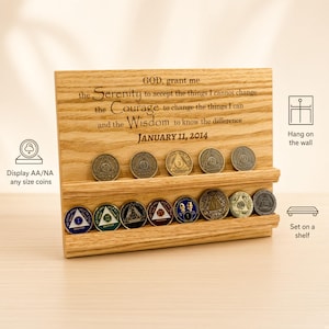 May include: A rectangular light-colored wooden display stand with two horizontal grooves for holding coins. The wood is engraved with the Serenity Prayer and the date January 11, 2014. Several circular metal recovery coins are placed in the grooves.