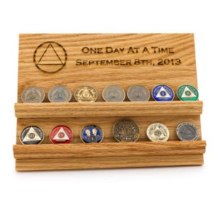 May include: A light brown wooden display stand with two horizontal shelves holding fourteen circular recovery coins. The top of the stand is engraved with a triangle symbol and the text One Day At A Time September 8th, 2013. The coins feature various colors, numbers, and symbols.