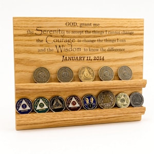May include: A light-colored wooden display stand featuring two horizontal grooves for holding circular tokens. The wood is engraved with the Serenity Prayer and the date January 11, 2014. Thirteen metallic recovery coins are arranged in the grooves.