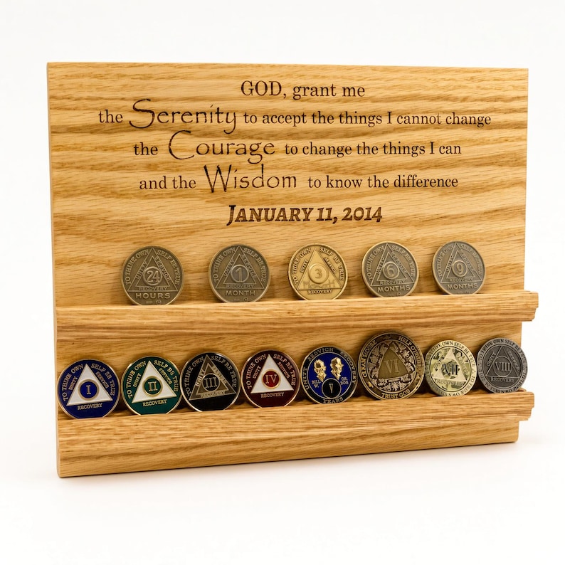 May include: A light-colored wooden display stand featuring two horizontal grooves for holding circular tokens. The wood is engraved with the Serenity Prayer and the date January 11, 2014. Thirteen metallic recovery coins are arranged in the grooves.