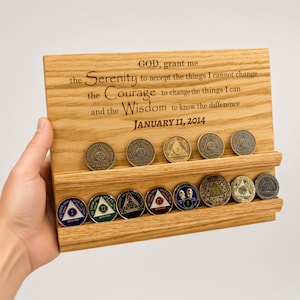 May include: A rectangular light-colored wooden display stand with two horizontal grooves holding fifteen circular metal sobriety coins. The wood is engraved with the text: GOD, grant me the Serenity to accept the things I cannot change the Courage to change the things I can and the Wisdom to know the difference JANUARY 11, 2014.