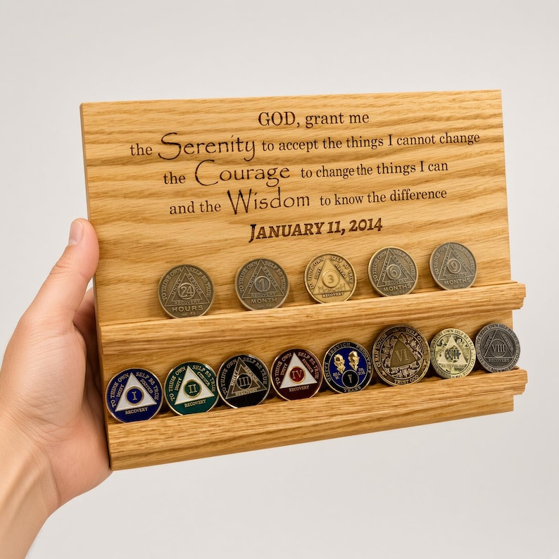 May include: A rectangular light-colored wooden display stand with two horizontal grooves holding fifteen circular metal sobriety coins. The wood is engraved with the text: GOD, grant me the Serenity to accept the things I cannot change the Courage to change the things I can and the Wisdom to know the difference JANUARY 11, 2014.