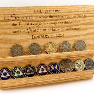 May include: A light-colored wooden display stand with two horizontal grooves holding twelve circular recovery coins. The wood is engraved with the Serenity Prayer and the date JANUARY 11, 2014. The coins feature various colors, numbers, and symbols.