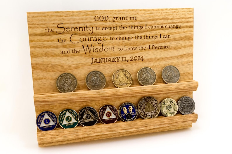 May include: A light-colored wooden display stand with two horizontal grooves holding twelve circular recovery coins. The wood is engraved with the Serenity Prayer and the date JANUARY 11, 2014. The coins feature various colors, numbers, and symbols.