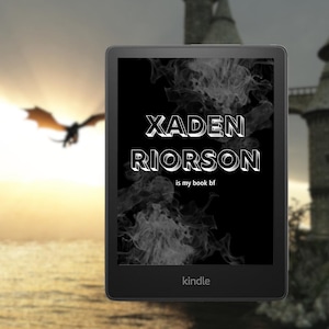 May include: A black and white Kindle e-reader with the text "Xaden Riorson is my book bf" on the screen. The background is a blurry image of a dragon flying in front of a castle.