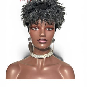 May include: A curly, salt-and-pepper grey wig styled with a black headband. The wig is displayed on a mannequin head wearing long, dark, oval-shaped drop earrings and a wide, silver-toned rhinestone choker necklace. The text WIGS by Caphy is visible.