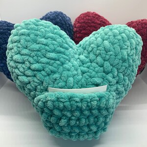 May include: A teal blue heart-shaped plush pillow with a white card tucked inside. The pillow is made of a soft, fuzzy material and has a textured surface.