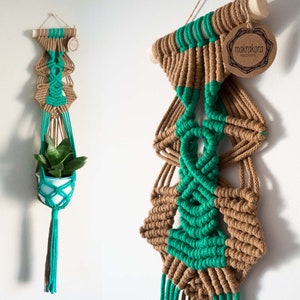 May include: A macrame plant hanger with a brown and teal color scheme. The hanger is made of knotted cord and has a wooden dowel at the top. The hanger is hanging from a white wall. The text "makrakora macrame" is on a brown tag.