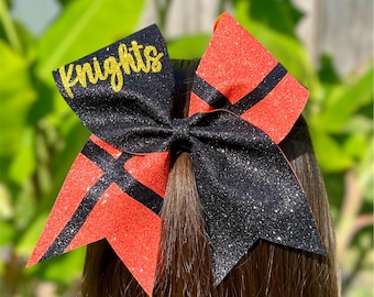 May include: A large hair bow featuring a glittery black and orange design with a split colour pattern. The left side is black with the word Knights written in gold glitter script, while the right side features orange and black diagonal stripes.